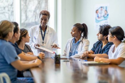 A small group of diverse nursing students sit around a boardroom table as they listen attentively to their teacher and lead doctor. They are each dressed in medical scrubs and sitting with papers out in front of them. The doctor is holding out a clipboard with a document on it as she reviews it with the group.